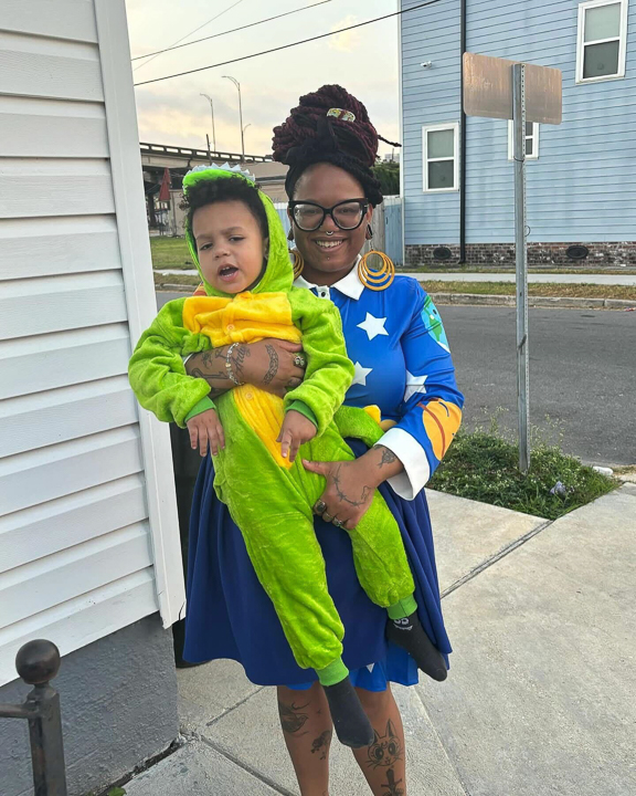 Ashley holding her child dressed in a dinosaur costume while wearing a colorful outfit outdoors