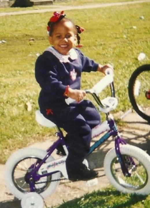 Young Ashley smiling while riding a small bicycle outdoors on a sunny day
