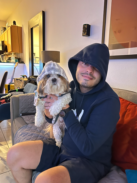 Renan sitting on a couch holding a small dog wearing a hoodie indoors